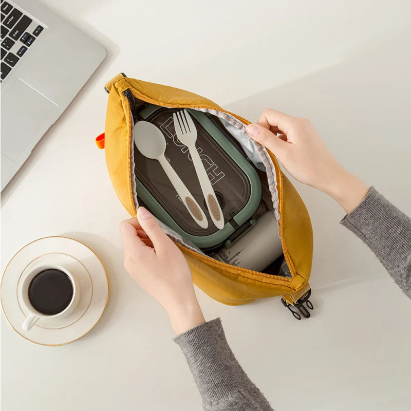 Person opening a yellow bag containing a lunch box, cutlery, and a mug on a desk with a laptop and coffee.